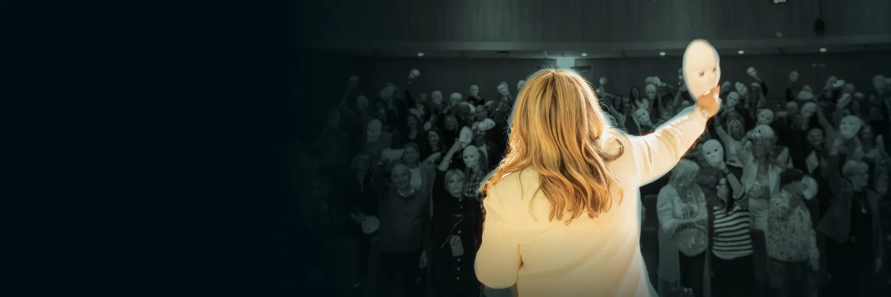 A speaker on stage holds a white mask up to an audience holding masks at the National Coaching Conference.