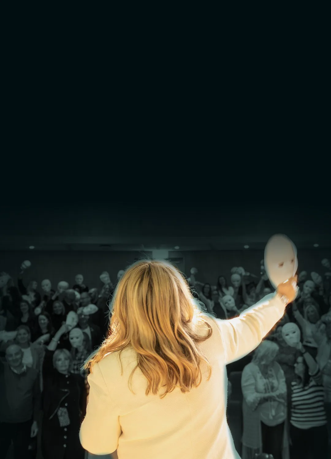A speaker on stage holds a white mask up to an audience holding masks at the National Coaching Conference.